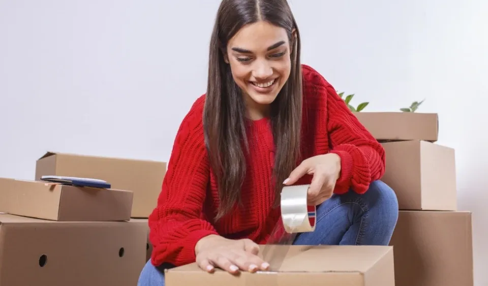 A woman, surrounded by moving boxes, tapes a box closed.